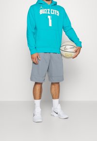 Felpa color teal con "BUZZ CITY" in bianco e un numero, pantaloni corti grigi, scarpe Nike bianche, mentre tiene un pallone da basket. Tessuto morbido, vestibilità atletica.