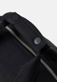 Sac à dos en nylon noir avec une texture lisse, doté d'une fermeture éclair et de boutons-pression noirs sur le dessus ainsi qu'une fermeture éclair argentée.