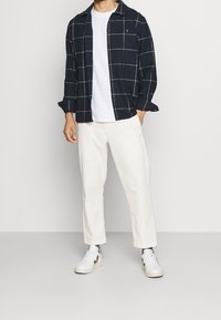 Camicia a quadri blu navy con dettagli bianchi, t-shirt bianca sotto, abbinata a pantaloni color crema e sneaker bianche.