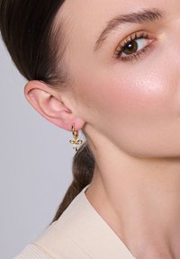 Złote kolczyki w formie kółek z małym akcentem w postaci kokardki. Gładkie metalowe wykończenie, lekka konstrukcja oraz wyrazisty wygląd zaprezentowane w zbliżeniu.