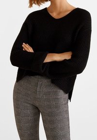 Czarny sweter z dzianiny, z dekoltem w serek i podwiniętymi rękawami, w zestawieniu z szarymi, w kratę spodniami o wysokim stanie z teksturowanym wzorem.