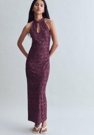 Femme portant une robe sans manches longue jusqu’au sol de couleur violet foncé à motif floral, avec un décolleté en trou de serrure et des sandales blanches à bouts ouverts, debout devant un fond uni.