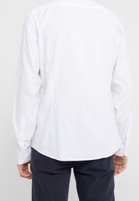 Henkilö, jolla on päällä pitkähihaisessa valkoinen paita ja tummat housut, näkyy takaa, seisahtuneena yksinkertaista vaaleaa taustaa vasten.