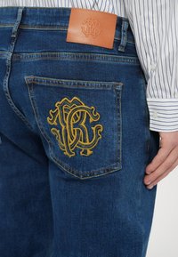 Vaqueros de mezclilla con un tono azul profundo, con un parche de cuero tostado y un logo bordado amarillo en el bolsillo trasero, detalles de costuras en contraste.