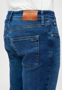 Τζιν παντελόνι σε σκούρο μπλε χρώμα με υφή, διαθέτοντας δερμάτινη ετικέτα με την ένδειξη "TOMMY JEANS" στη μέση και ορατές πίσω τσέπες.