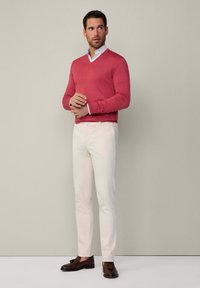 Pullover aus rosa Strick mit V-Ausschnitt, kombiniert mit cremefarbenen Slim-Fit-Hosen und braunen Slippern mit Quasten. Minimalistischer Hintergrund.