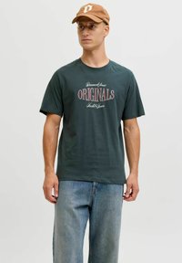 Camiseta de algodón verde oscuro con la palabra "ORIGINALS" en texto rojo y blanco. Combinada con unos vaqueros de mezclilla azul claro y una gorra marrón.