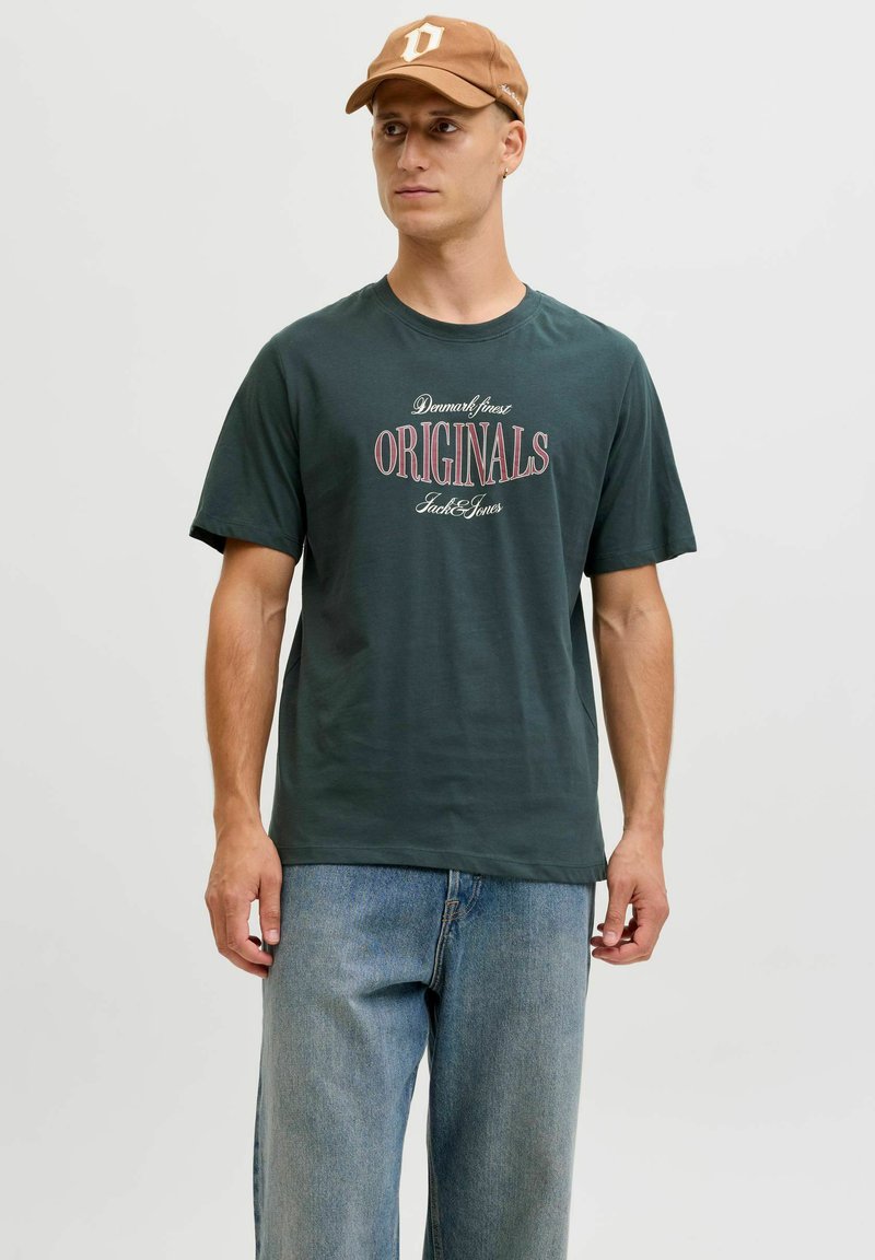 Camiseta de algodón verde oscuro con la palabra "ORIGINALS" en texto rojo y blanco. Combinada con unos vaqueros de mezclilla azul claro y una gorra marrón.