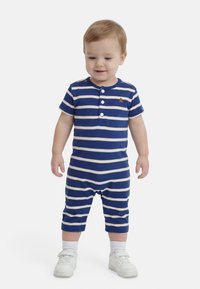 Νήπιο που στέκεται, φορώντας ένα ριγέ ρομπέρ με μπλε και λευκές ρίγες με κουμπιά και λευκά αθλητικά παπούτσια, μπροστά σε απλό λευκό φόντο.