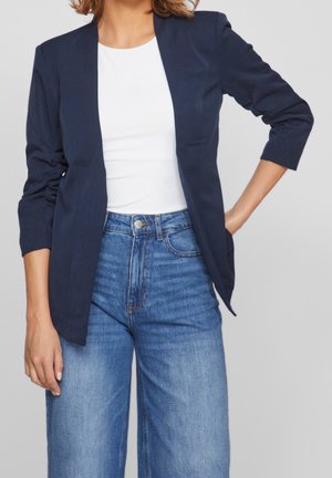 Person iført en navy blazer med trekvartærmer over en hvid top og højtaljede blå jeans, stående med den ene hånd på hoften.