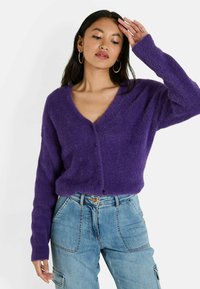 Cárdigan morado esponjoso con escote en V, cierre de botones en la parte frontal y mangas largas, combinado con unos vaqueros de cintura alta en azul claro.