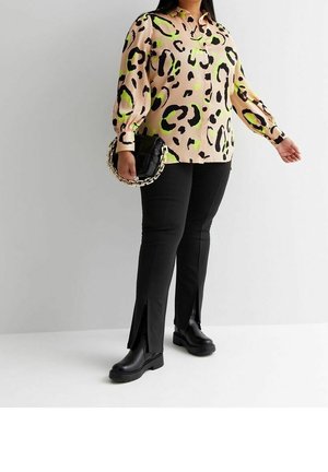 Femme portant un chemisier beige à imprimé léopard noir et vert, un pantalon noir fendu à l'ourlet, des bottes noires, et tenant un sac noir avec une chaîne dorée.