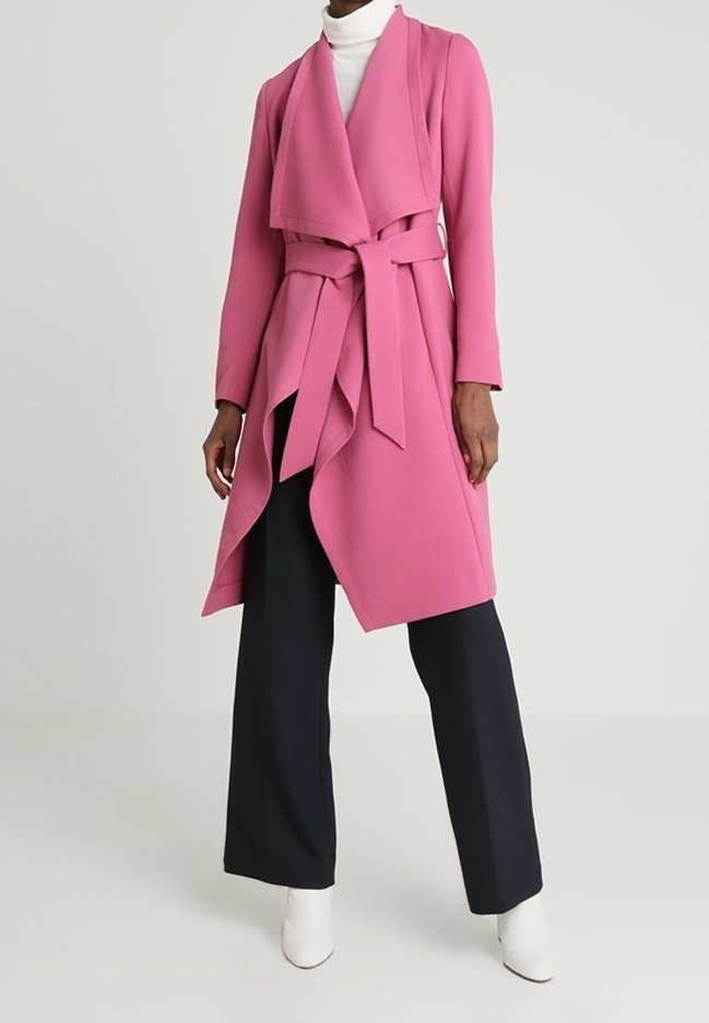Rosa Wickeljacke aus weichem Stoff, mit drapiertem Vorderteil, langen Ärmeln und einem Stoffgürtel; kombiniert mit marineblauen weit geschnittenen Hosen und weißen Stiefeletten.