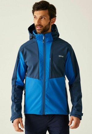 Mężczyzna w niebiesko-granatowej kurtce z kapturem i kieszeniami na zamek, stojący na jasnym tle, patrzący w bok.