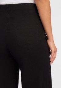 Zwarte broeken met een gladde textuur, hoge taille en een enkele achterzak met een gevormde klep, weergegeven vanuit een zijaanzicht.