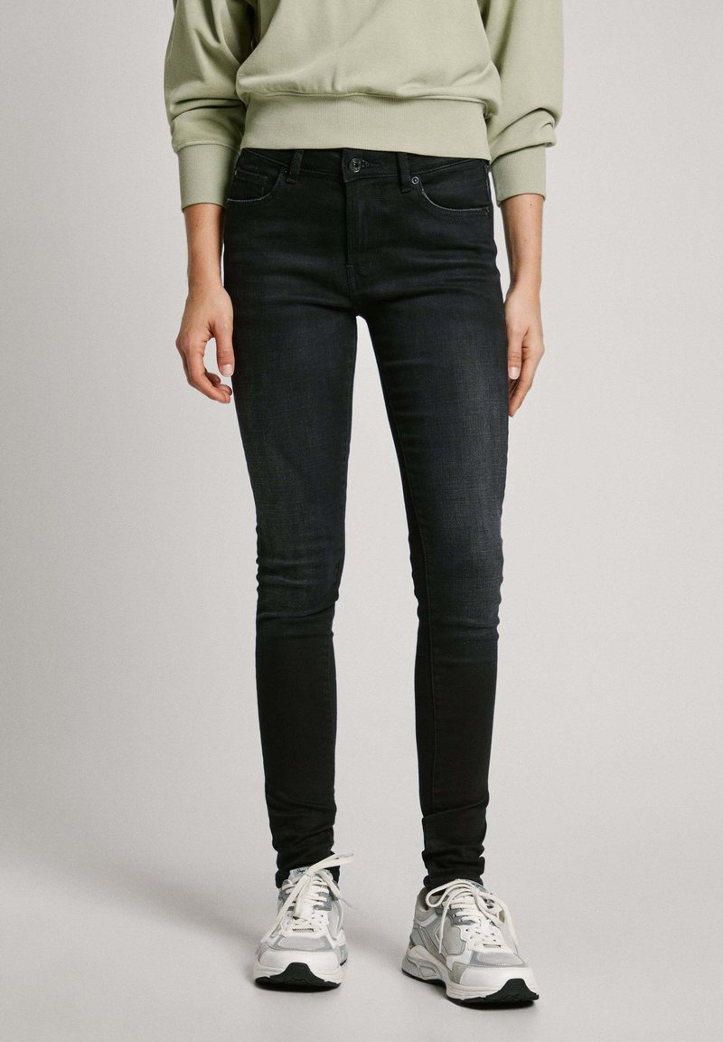 Juodi liesi džinsai, pagaminti iš džinsinio audinio, su klasikinio penkių kišenių dizainu ir subtiliais siuvinėjimo akcentais. Dėvimi su pilkais sportiniais batais.