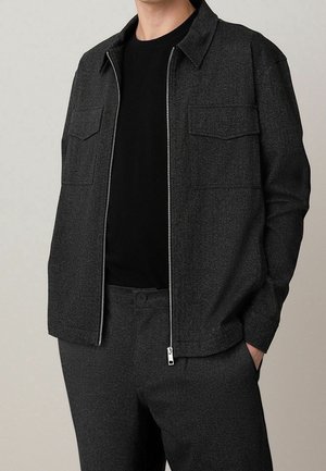 Mężczyzna ubrany w ciemnoszarą kurtkę zapinaną na zamek z klapami na kieszeniach na piersi, na czarnej koszulce i pasujące ciemnoszare spodnie, z rękami w kieszeniach.