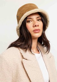 Bruine suède emmerierdop met een zachte crèmekleurige fleece rand. Het model draagt een lichtbeige textuurjack over een witte blouse.