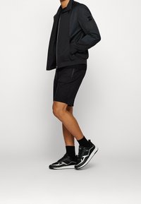 Homme portant une veste noire zippée, un short noir, des chaussettes noires et des baskets noir et blanc, debout devant un fond gris clair.