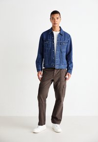 Donkerblauwe denimjas met knoopsluiting aan de voorkant, twee borstzakken en lange mouwen, gecombineerd met bruine cargobroek en witte sneakers.