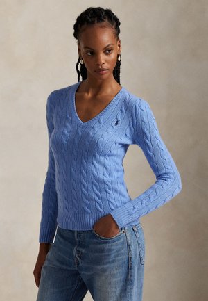 Donna con capelli intrecciati che indossa un maglione celeste a collo a V lavorato a trecce e jeans blu, posando con una mano in tasca.