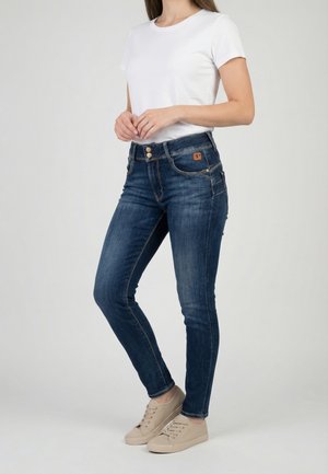 Femme portant un jean slim bleu foncé, un t-shirt blanc à manches courtes et des baskets beige, debout devant un fond gris uni.