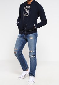 Uomo in giacca navy, t-shirt grafica navy, jeans blu strappati e sneaks bianche, in piedi con le mani nelle tasche.