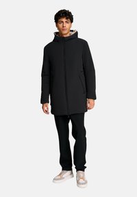 Uomo che indossa un cappotto nero con cappuccio, pantaloni neri e sneakers bianche con accenti arancioni, in piedi contro uno sfondo chiaro e semplice.