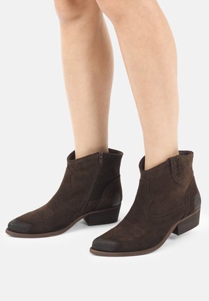 Brązowe zamszowe botki za kostkę z niskimi, warstwowymi obcasami i bocznymi zamkami, noszone na gołych nogach na jednolitym tle.