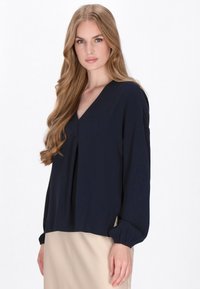 Marineblå bluse i let stof med V-hals, løs pasform og ballonærmer. Vist med en beige nederdel. Enkel, blød tekstur.