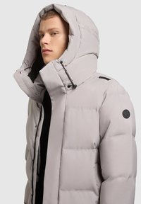 Gai pelēks pildīts parka ar lielu kapuci, melnu rāvējslēdzēju un sānu pogām. Iespējojiet modernu auduma tekstūru un apļveida logo uz pleca.