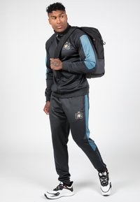 Zwarte sportieve trainingsoutfit met blauwe accenten, bestaande uit een zip-up jack en broek met elastische tailleband. Inclusief een zwarte rugzak.