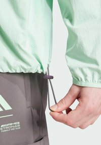 Miętowo-zielona lekka kurtka z elastycznym sznurkiem w talii, zestawiona z szarymi spodniami cargo z akcentami logo.