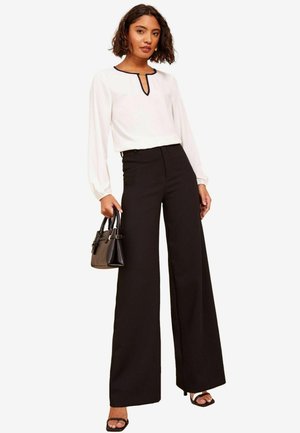 Blusa bianca a maniche lunghe con scollatura nera, abbinata a pantaloni neri a vita alta con gamba ampia. Gli accessori includono una piccola borsa nera.