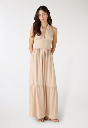Donna con capelli castani lunghi che indossa un abito lungo beige senza maniche con collo all'americana e sandali bassi, in piedi contro uno sfondo bianco semplice.