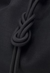 Sac en tissu noir doté de cordes noires robustes, finement nouées pour accentuer le design. La texture est lisse avec une finition mate.