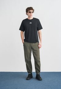 Μαύρο κοντομάνικο T-shirt με λογότυπο, συνδυασμένο με λαδί στενά παντελόνια. Το μοντέλο φοράει γυαλιά ηλίου και μαύρα αθλητικά παπούτσια. Ουδέτερο φόντο.