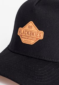 Mustra mustrikatega must müts, millel on pruun nahkpatch, mis on kujundatud rombiks, millel on reljeefselt kiri "BLACKSKIES PREMIUM SUPPLIES MMIX."