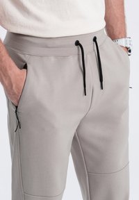 Joggers gris claro fabricados con una tela suave, presentan una cintura elástica con un cordón negro, bolsillos laterales y detalles con cremallera.