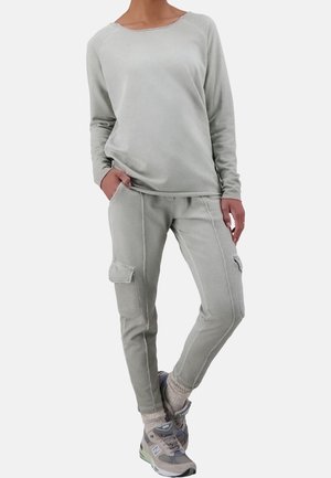 Frau trägt ein hellgraues Langarmshirt und passende Cargohosen mit Sneakern, steht mit einer Hand in der Tasche vor einem einfarbigen Hintergrund.