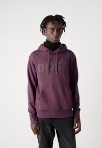 Lila Kapuzenpullover mit Fronttasche und Kordelzügen, mit großem "BOSS"-Logo in einem dunkleren Farbton. Über einem schwarzen Rollkragenpullover getragen.