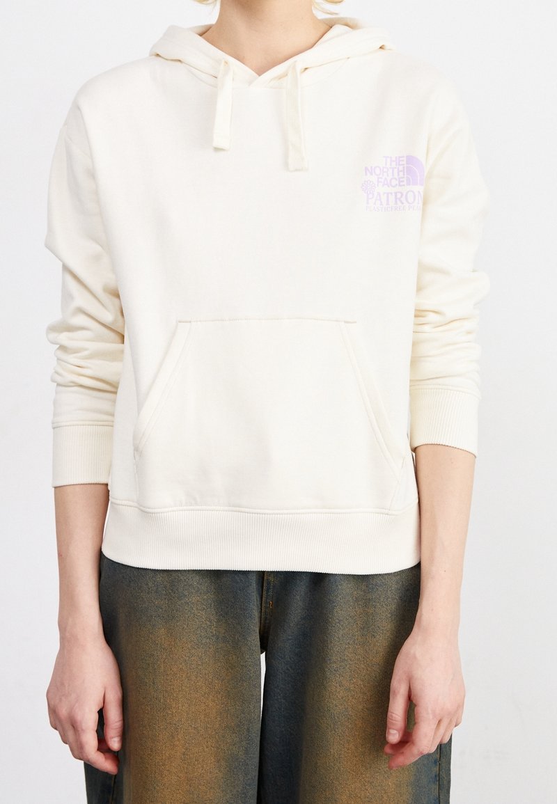 Personne portant un sweat à capuche crème avec cordons et poche avant, associé à un jean foncé délavé. Torse et bras partiellement visibles.