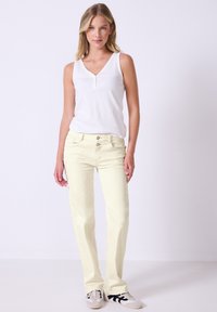 Jeans rectos color amarillo claro con cinco bolsillos y cierre de botón, combinados con una blusa sin mangas blanca y zapatillas con patrones.