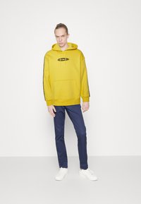 Yellow hoodie with front pocket and black logo, paired with navy trousers and white sneakers. Smooth texture and casual fit.