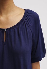 Marinblå topp med rundad halsringning och rynkdetalj. Har en liten knapp/flikstängning framtill, korta ärmar och mjukt material.