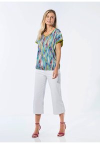Mehrfarbige Bluse mit abstrakten Streifen, grünen Akzenten und einem Rundhalsausschnitt, kombiniert mit weißen, knöchelfreien Hosen und fuchsiafarbenen Absatzsandalen.