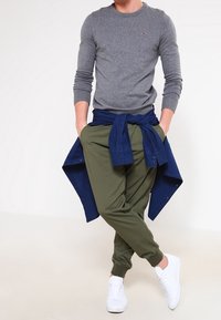 Šedé tričko s dlouhým rukávem, olivově zelené tepláky a bílé tenisky. Tmavě modré tričko uvázané kolem pasu. Ležérní střih s žebrovanými manžetami.