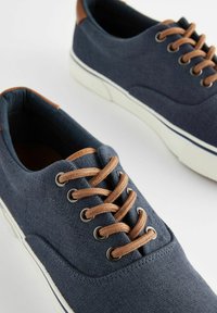 Marine canvas sneakers met bruine leren accenten, ronde neus en een contrasterende witte rubberen zool. Beschikt over bruine veters en metalen oogjes.