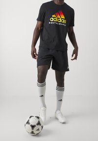 Mężczyzna ubrany w czarną koszulkę Adidas Deutschland, czarne szorty, białe skarpety do kolan Adidas oraz białe korki z czarno-białą piłką nożną Adidas.