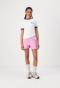 Hvit T-skjorte med marineblå kant og logo, sammen med rosa shorts og hvite knestrømper. Fottøyet er lyserosa joggesko med svarte detaljer.